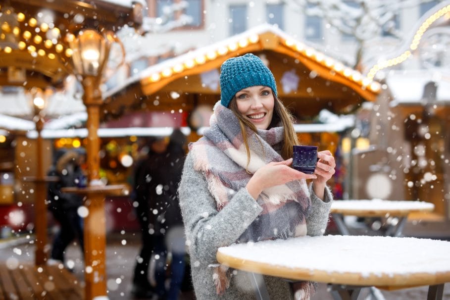 Beautiful young woman drinking hot punch, mulled wine on German Christmas market. Happy girl in winter clothes with lights on background on winter snow day in Dresden, Germany