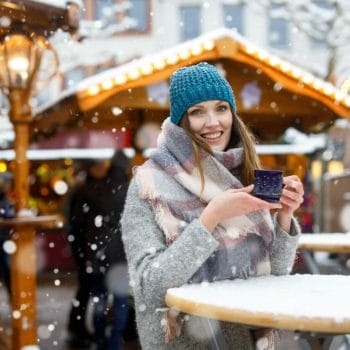 Beautiful young woman drinking hot punch, mulled wine on German Christmas market. Happy girl in winter clothes with lights on background on winter snow day in Dresden, Germany