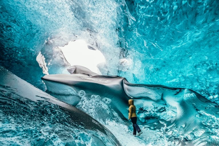 Woman wearing a yellow jacket stood inside a blue glacier ice cave.
