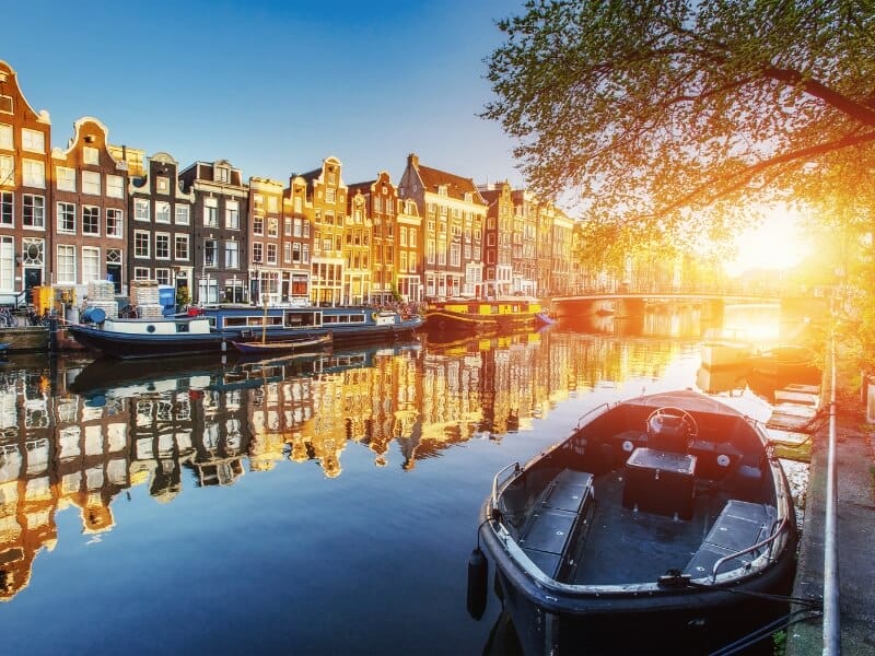 View of the Amsterdam canals during sunset. Photo shows the canal houses on the left with canal boats lining the sides of the canals.