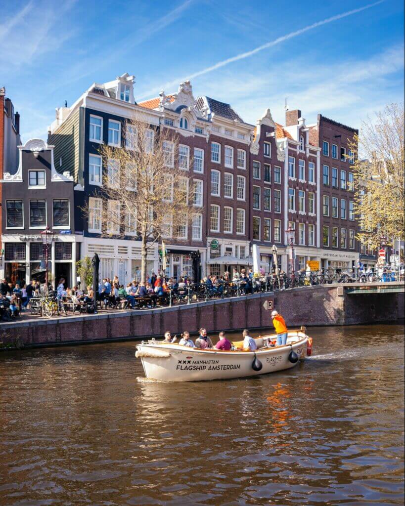 An open-top Flagship Amsterdam boat, which is white with the words "Manhatten, Flagship Amsterdam" written on the side cruising along the canals in Amsterdam. 