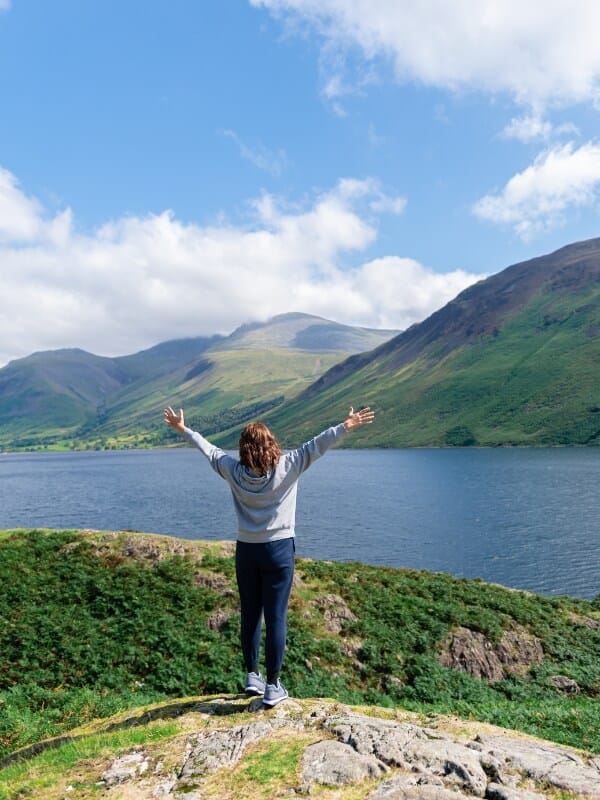 Woman stood in front of Waswater in the Lake District with brown hair, wearing a grey jumper and jeans.
